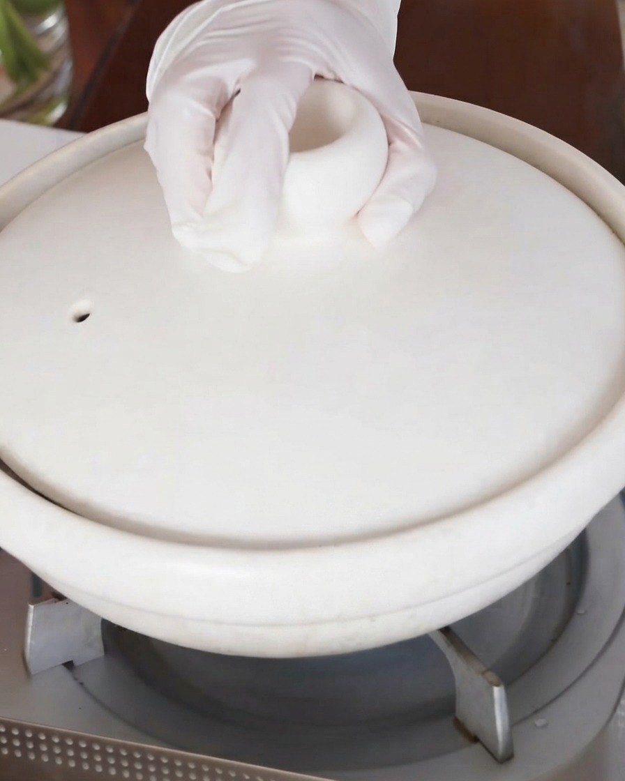 A hand placing the lid onto the white ceramic braising pot on the stove.