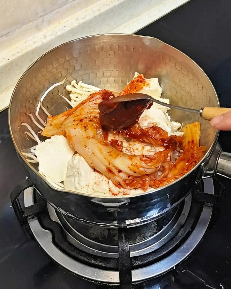 A spoon adding a dollop of thick red Korean chili paste onto the ingredients in the pot.