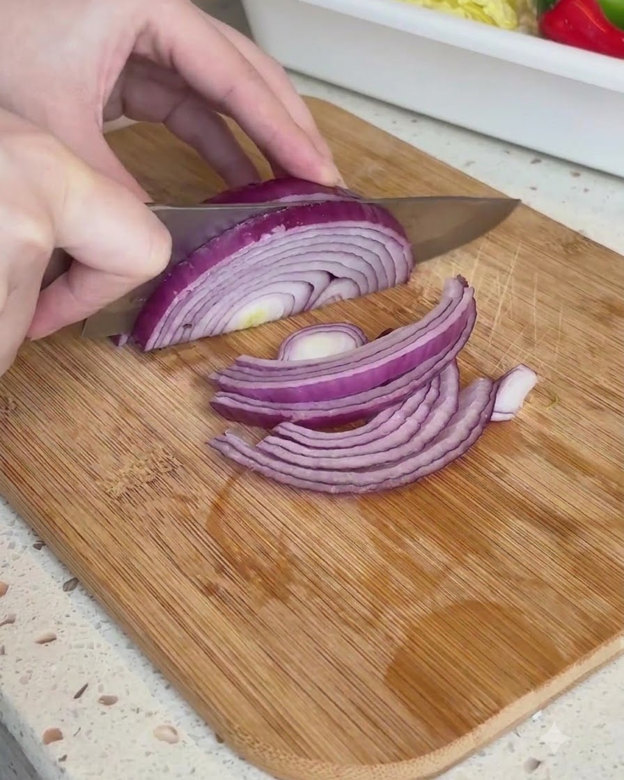 Half of a red onion being cut into thin strips on a wooden cutting board.