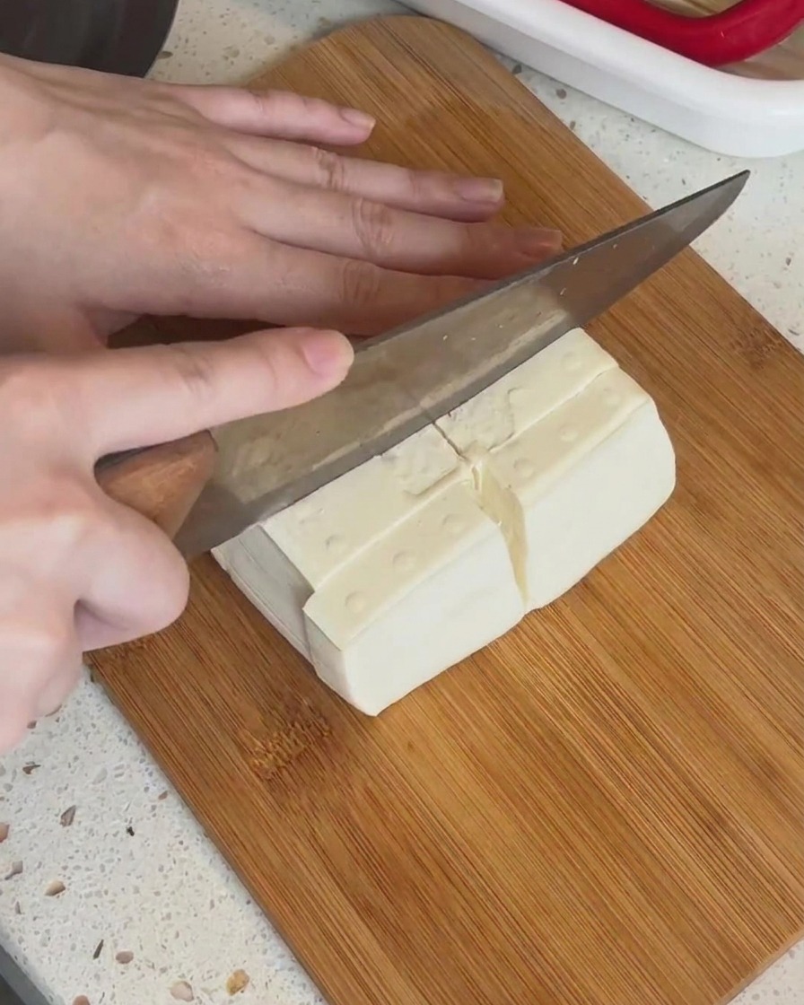 A block of white tofu being sliced into smaller rectangular pieces on a wooden board.