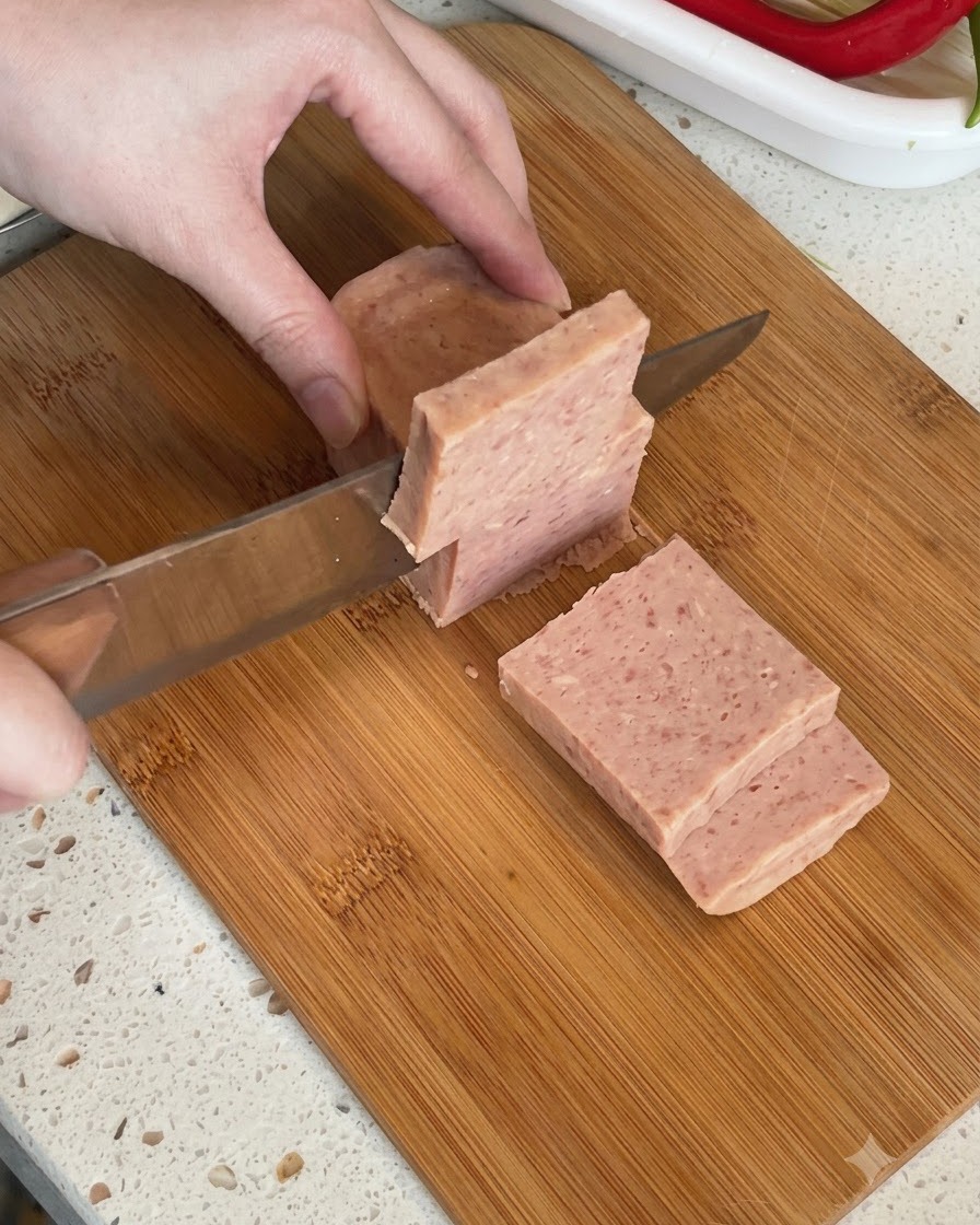 A block of canned luncheon meat being sliced into thick pieces on a cutting board.