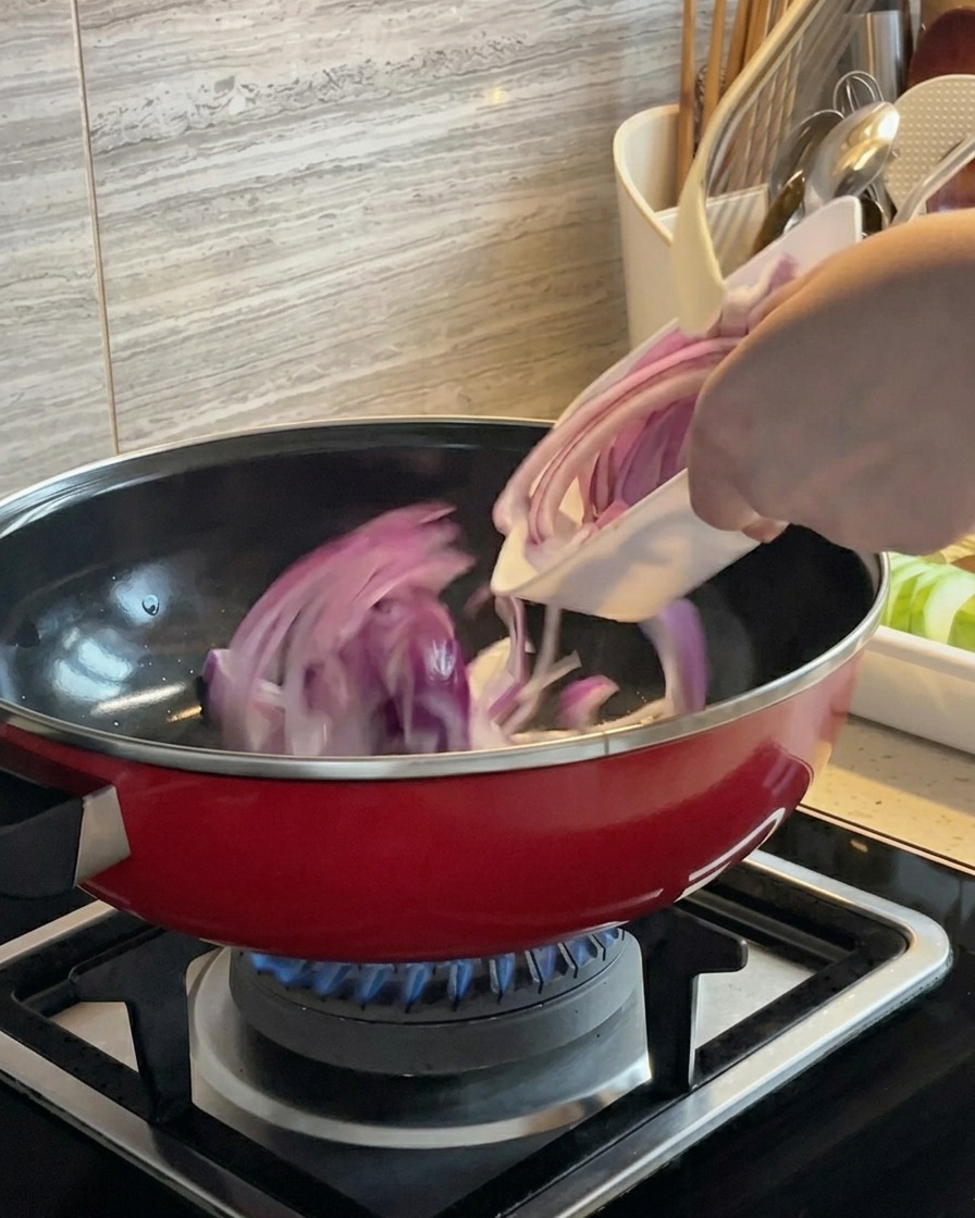 Slices of red onion being tossed and stir-fried in a red pot over a gas burner.
