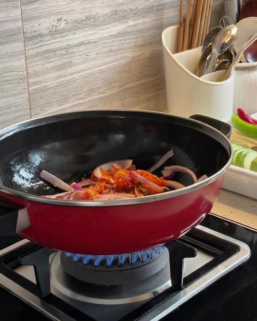 Sliced red onions and chopped kimchi being stir-fried together in a red wok over a blue flame.