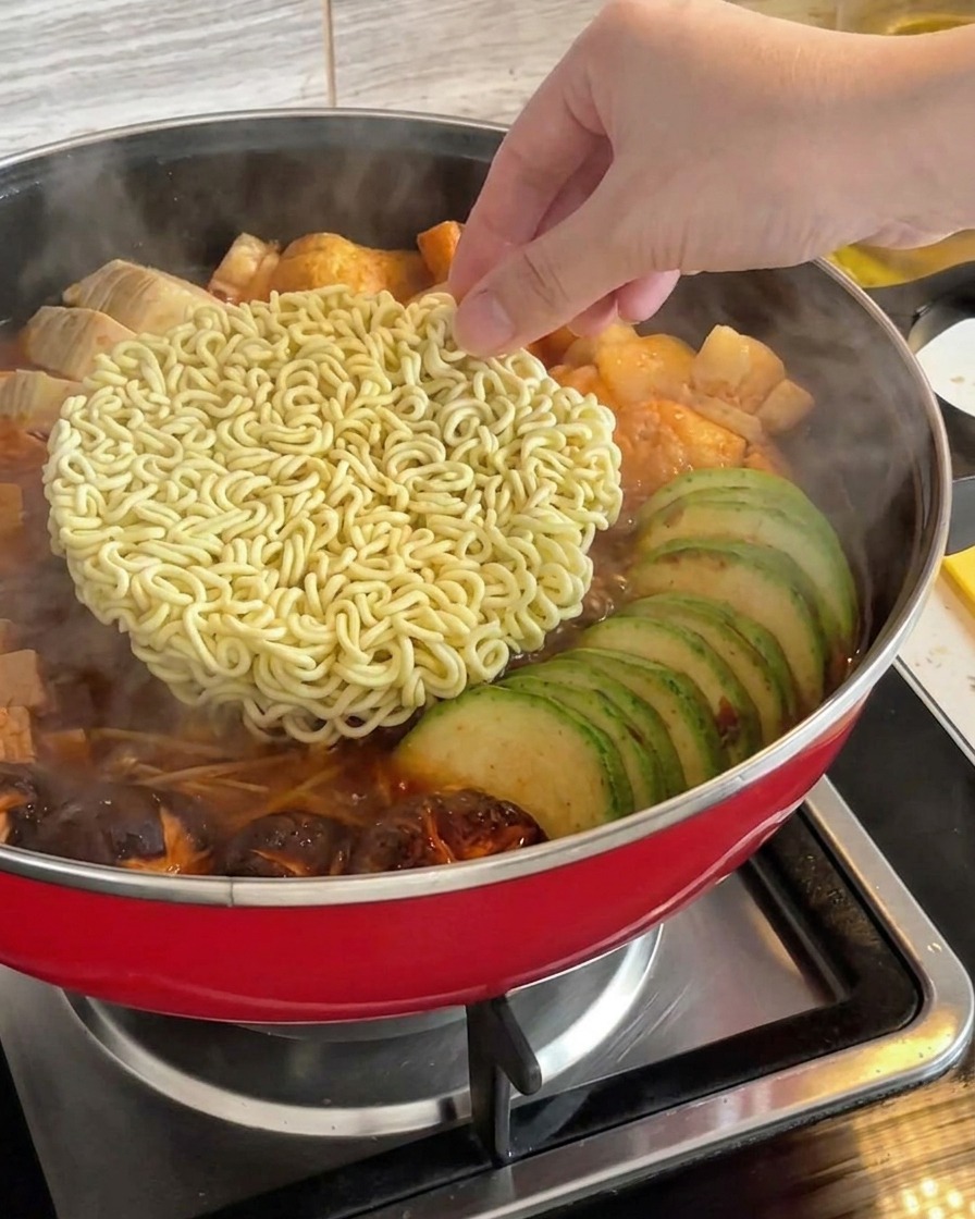 A hand placing a block of dry instant ramen noodles into the center of a simmering Korean army stew.
