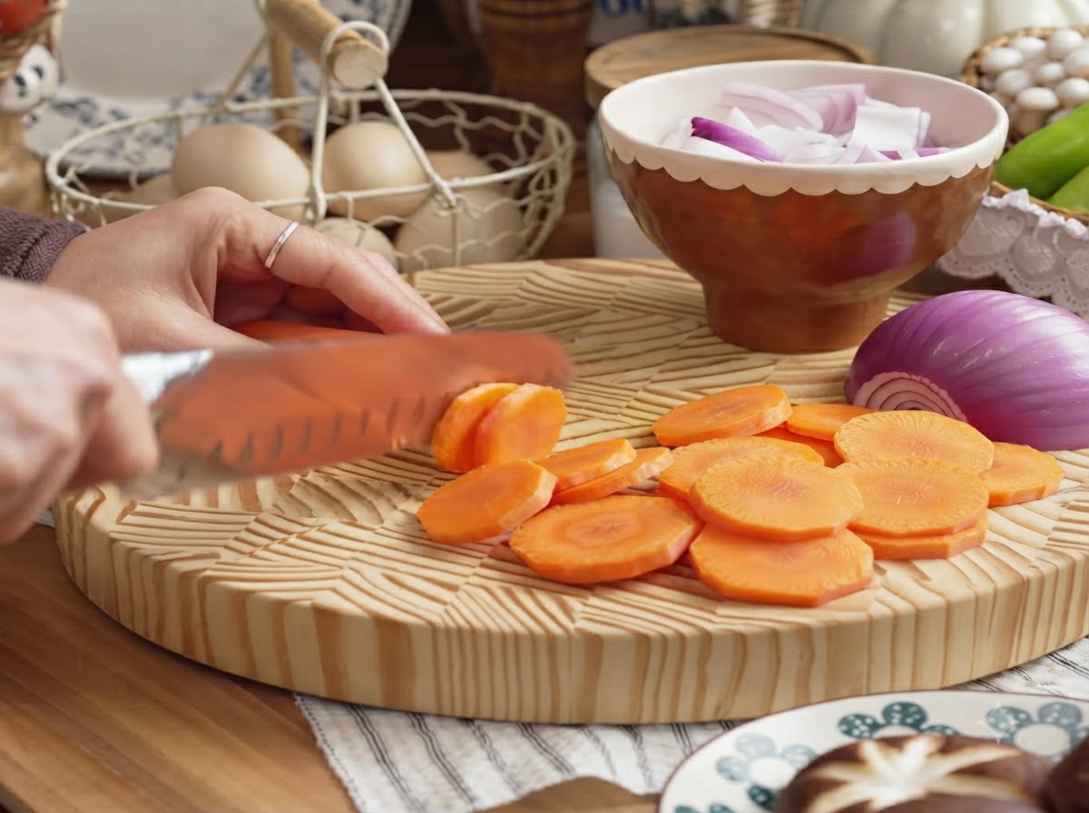 A hand slicing a peeled carrot into round coins on a wooden cutting board.
