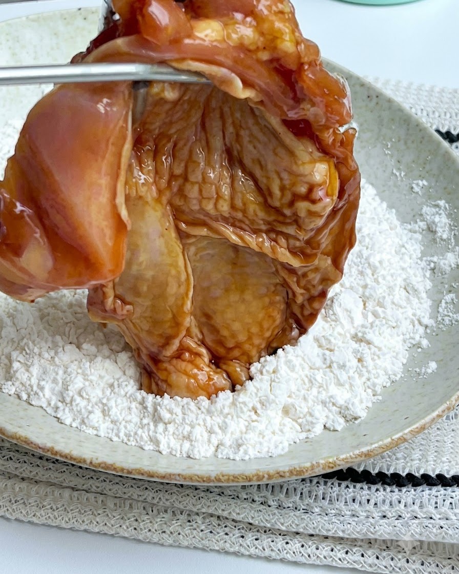Tongs holding a marinated chicken piece over a white plate filled with white cornstarch powder.
