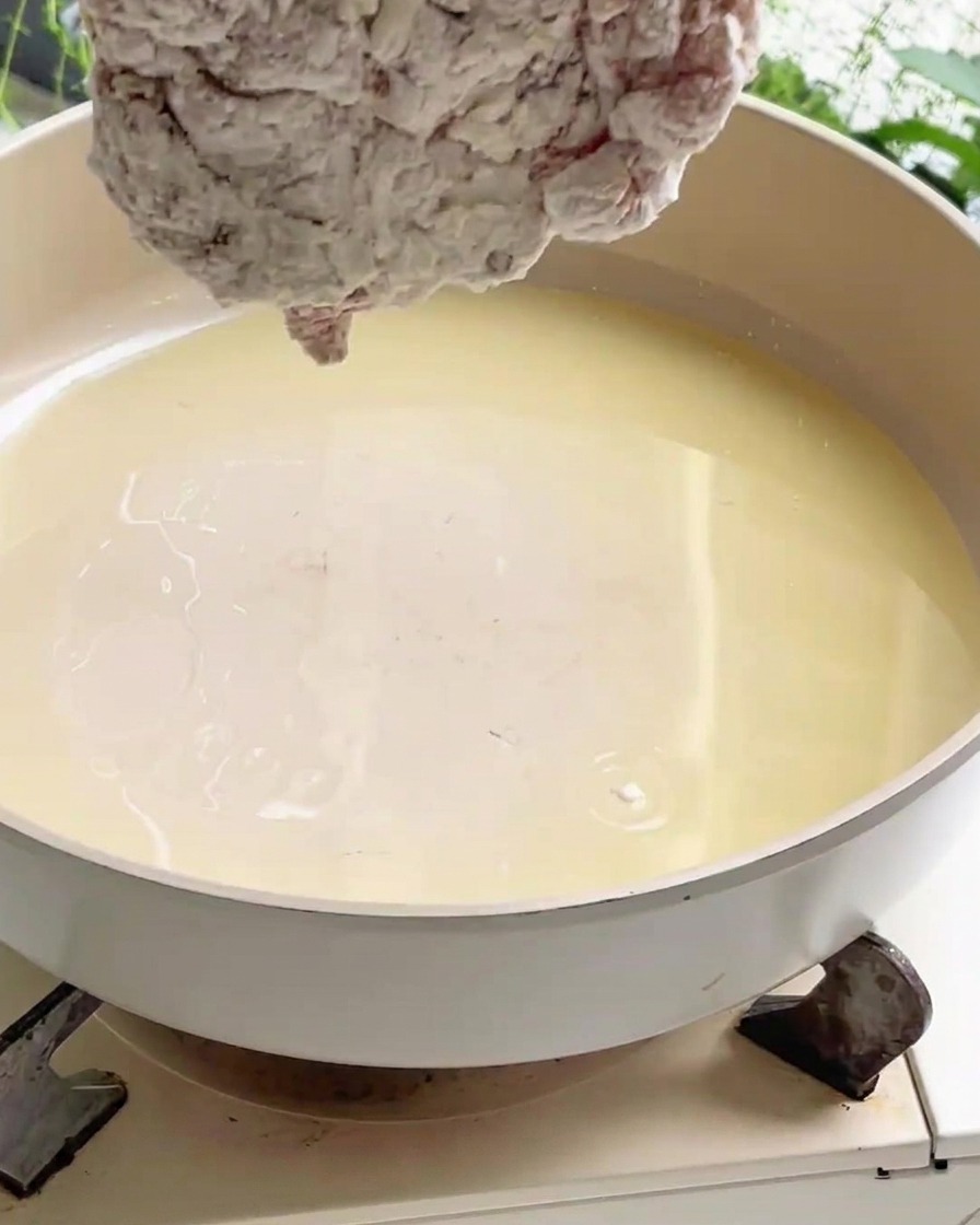A piece of coated chicken being lowered into a wide pan filled with hot cooking oil on a stove.