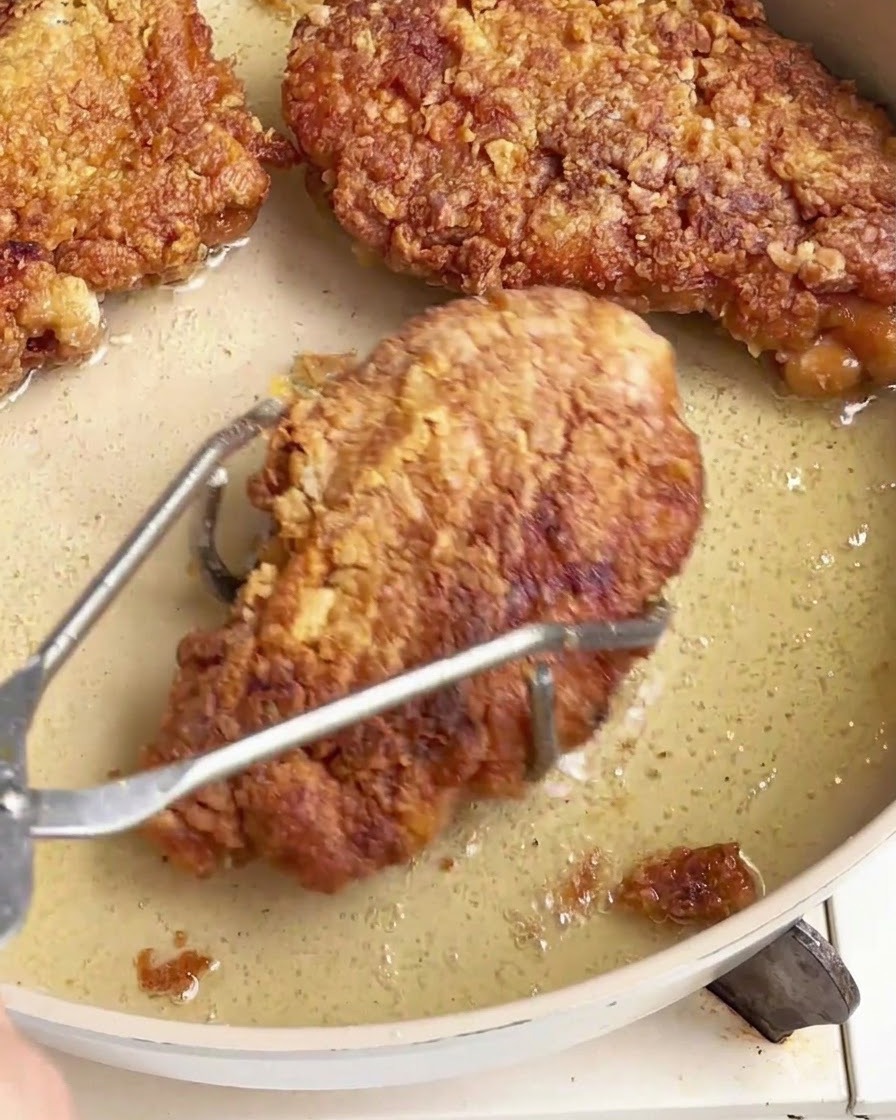 Crispy fried chicken chops being lifted out of an oily pan using stainless steel tongs.