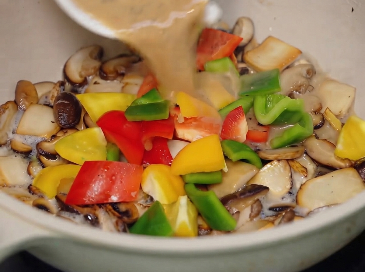 The black pepper sauce being poured from a small white bowl into the pan containing mixed mushrooms and colorful bell peppers.