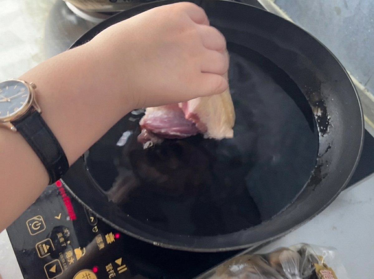 A hand placing a piece of pork into a pan of cold water with ginger and cooking wine.
