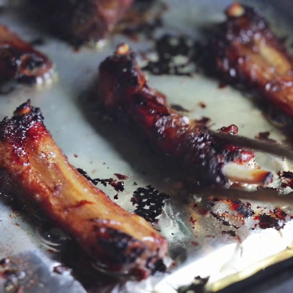 Close-up of grilled pork ribs being turned over on a foil-lined baking sheet with chopsticks.