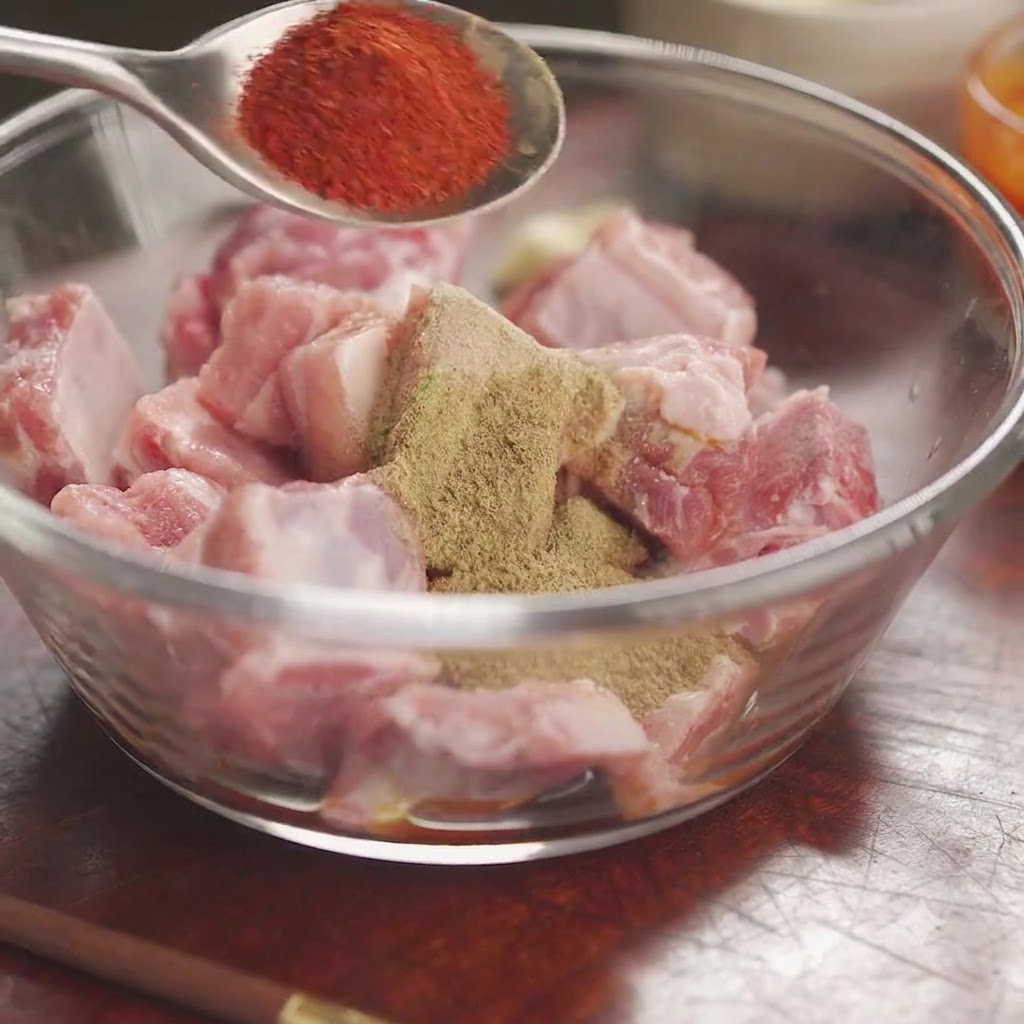Red chili powder on a spoon held over a bowl of raw pork ribs covered with mixed spices and herbs.