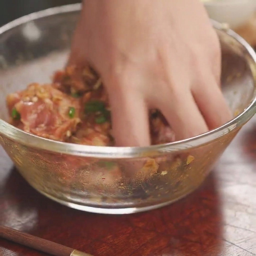 A bare hand thoroughly mixing seasoned raw pork ribs inside a clear glass bowl.