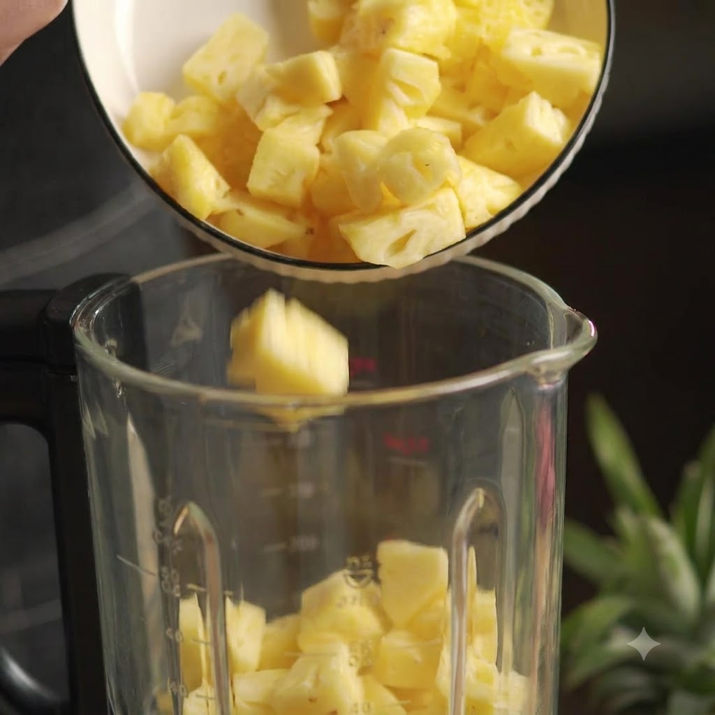 Fresh diced pineapple pieces being poured from a white bowl into a glass blender.