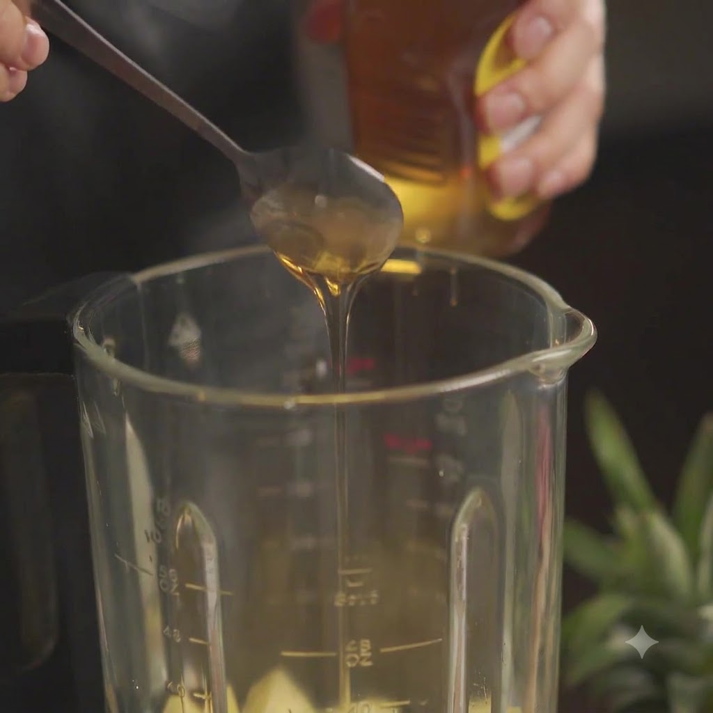 A spoonful of honey being added into a blender jar filled with chopped pineapple.