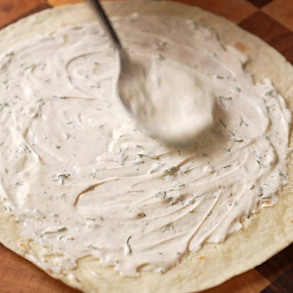 Dill sour cream sauce being spread evenly onto a wheat wrap with a spoon.
