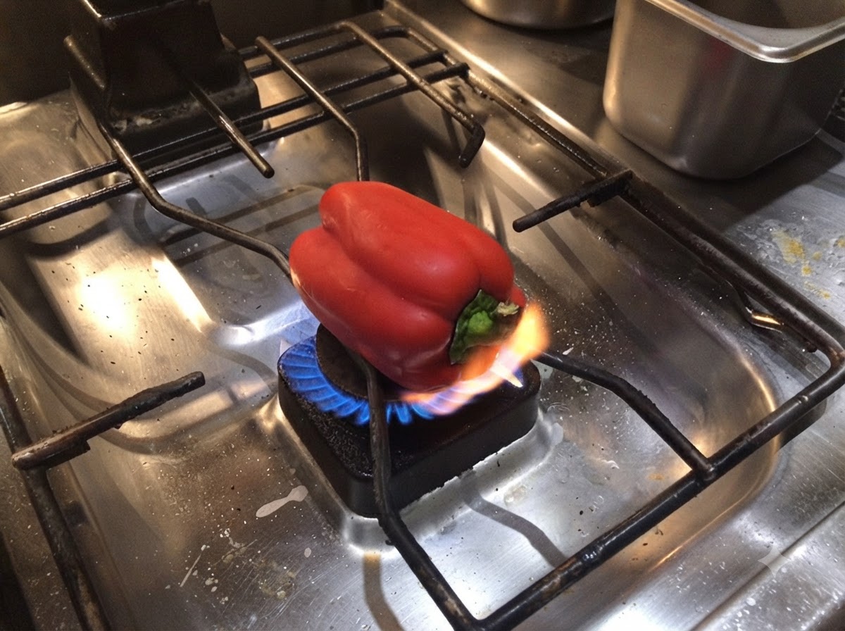A red bell pepper resting on a gas stove burner with an open flame charring its base.