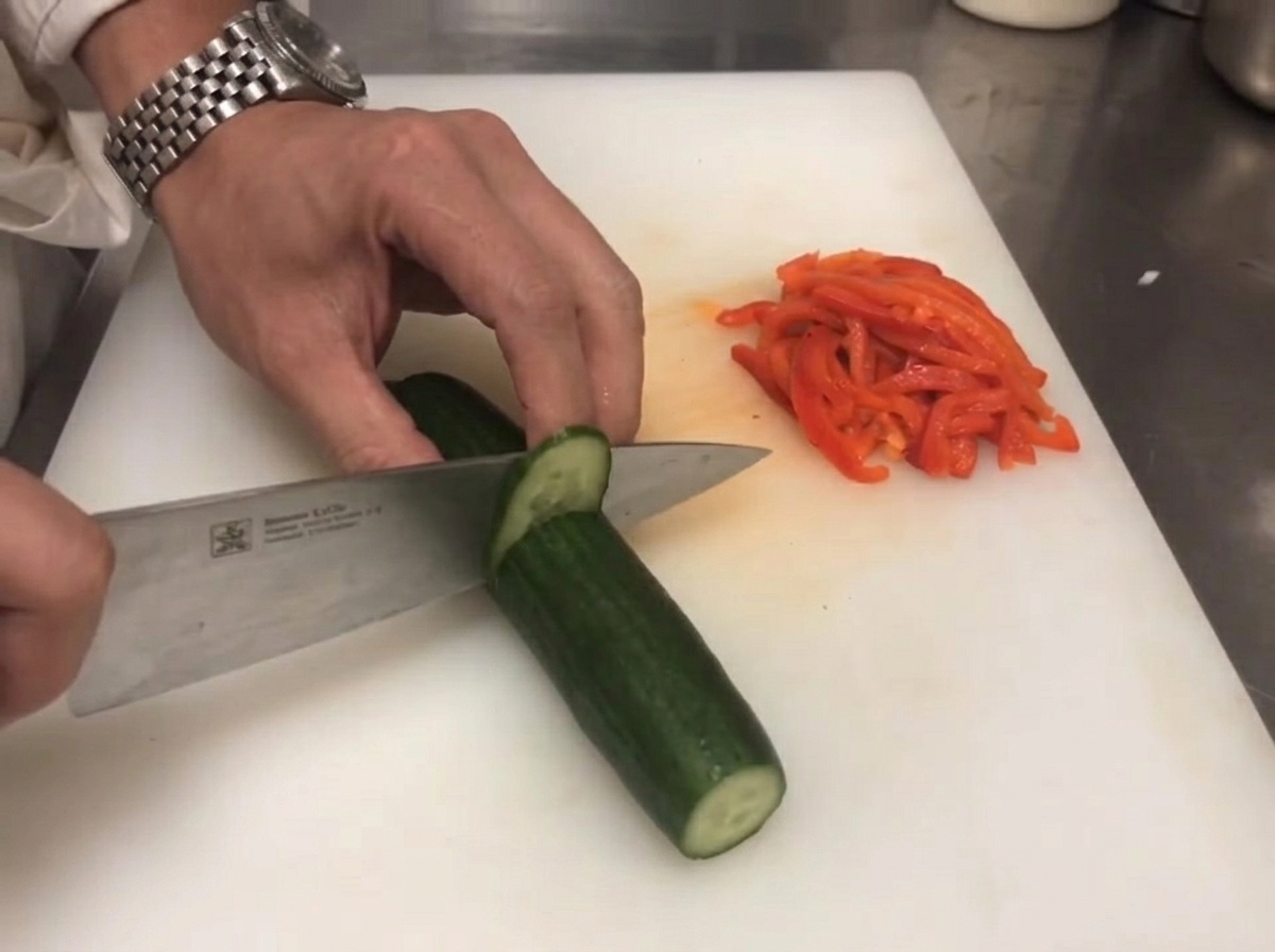 A knife slicing through a fresh green cucumber next to sliced red bell peppers.