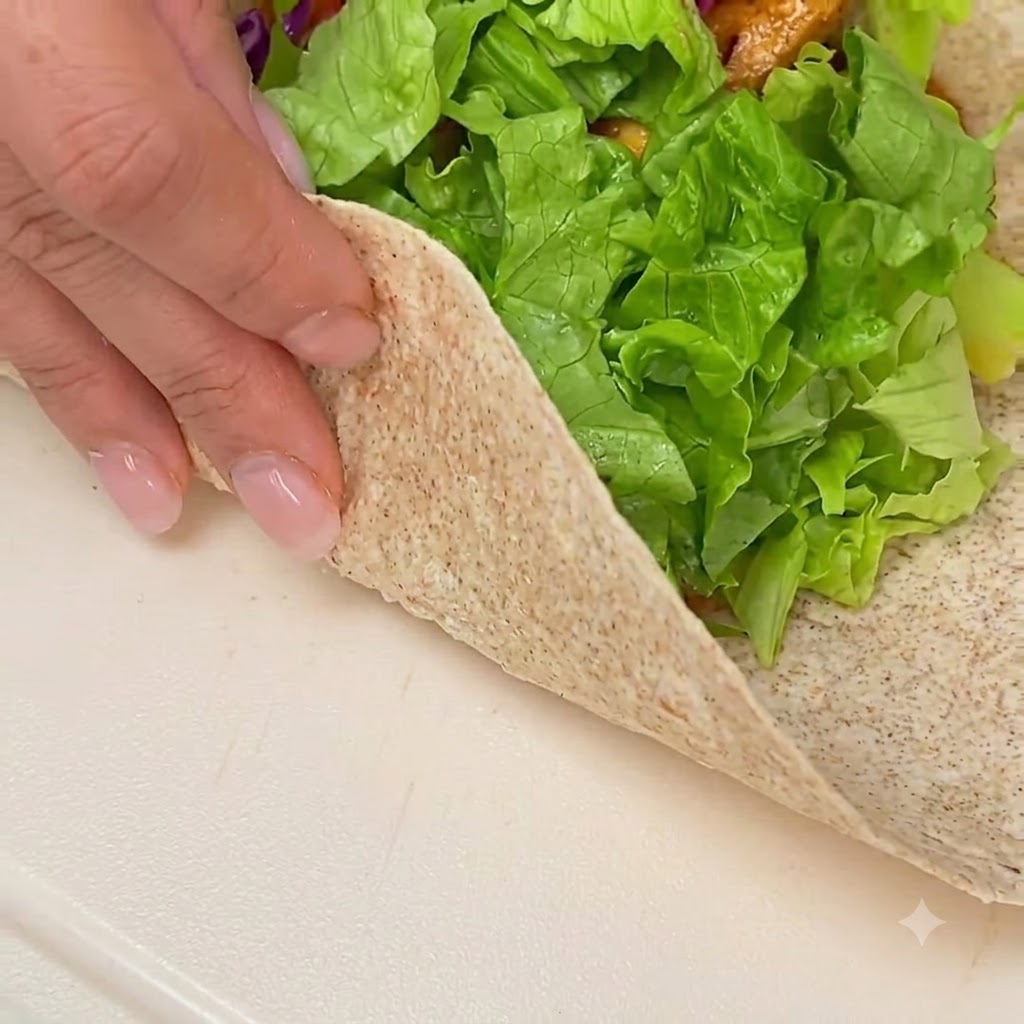 Hands folding and rolling a stuffed whole wheat tortilla on a clean white surface.