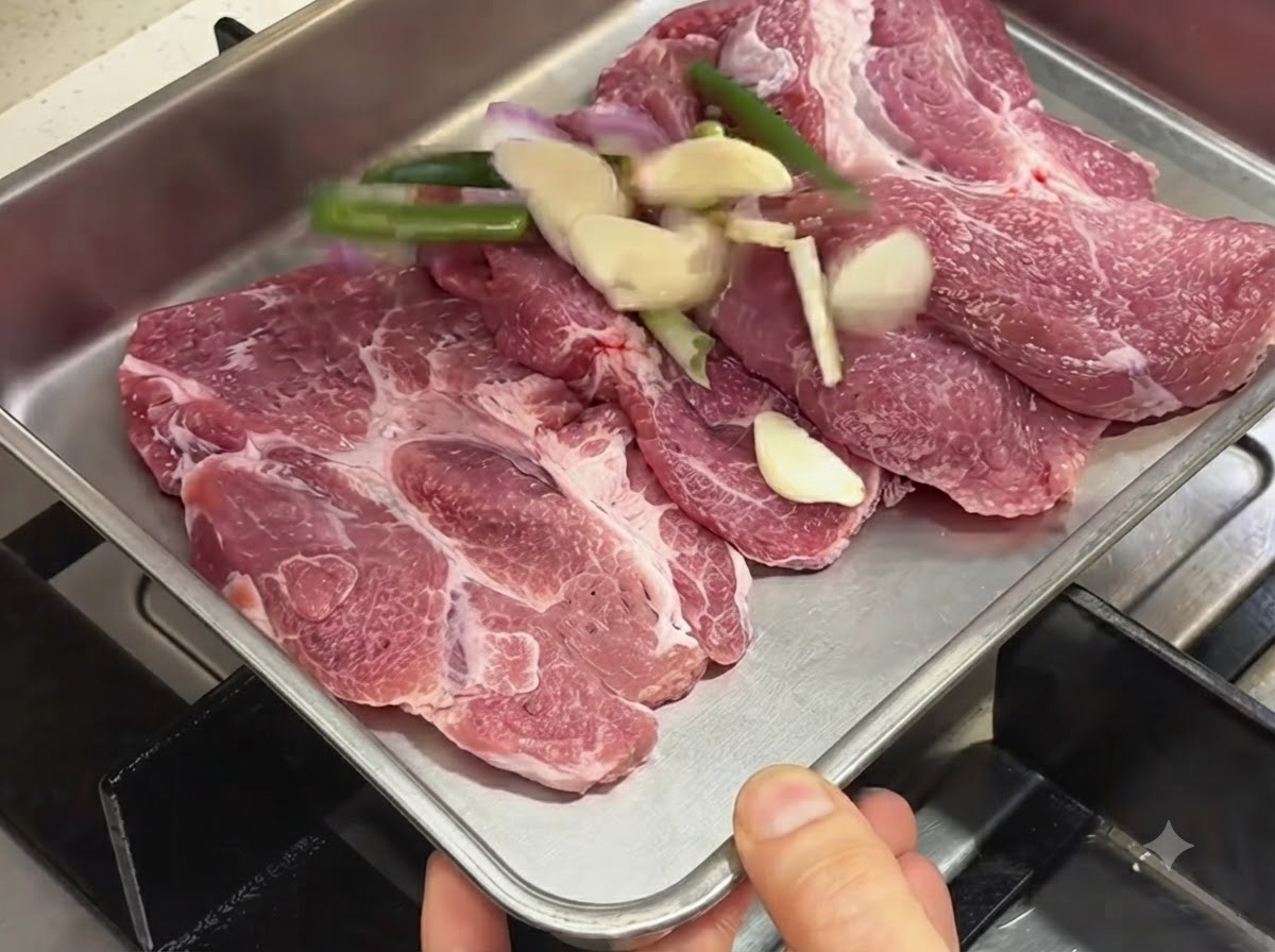 Raw pork chops arranged in a metal tray alongside sliced garlic, green onions, and shallots.