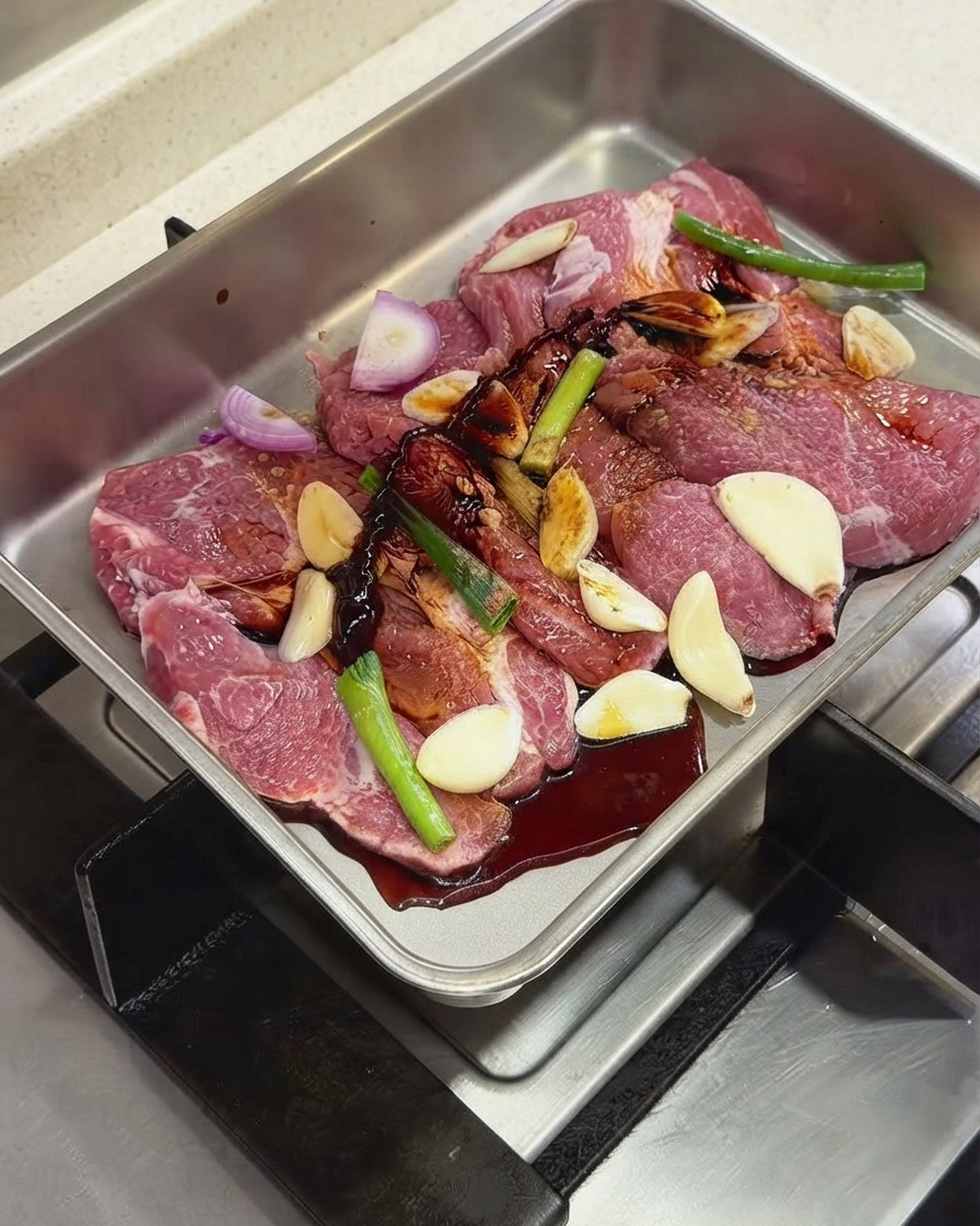 Dark sauces and seasonings being poured over the pork chops and aromatics inside a metal tray.