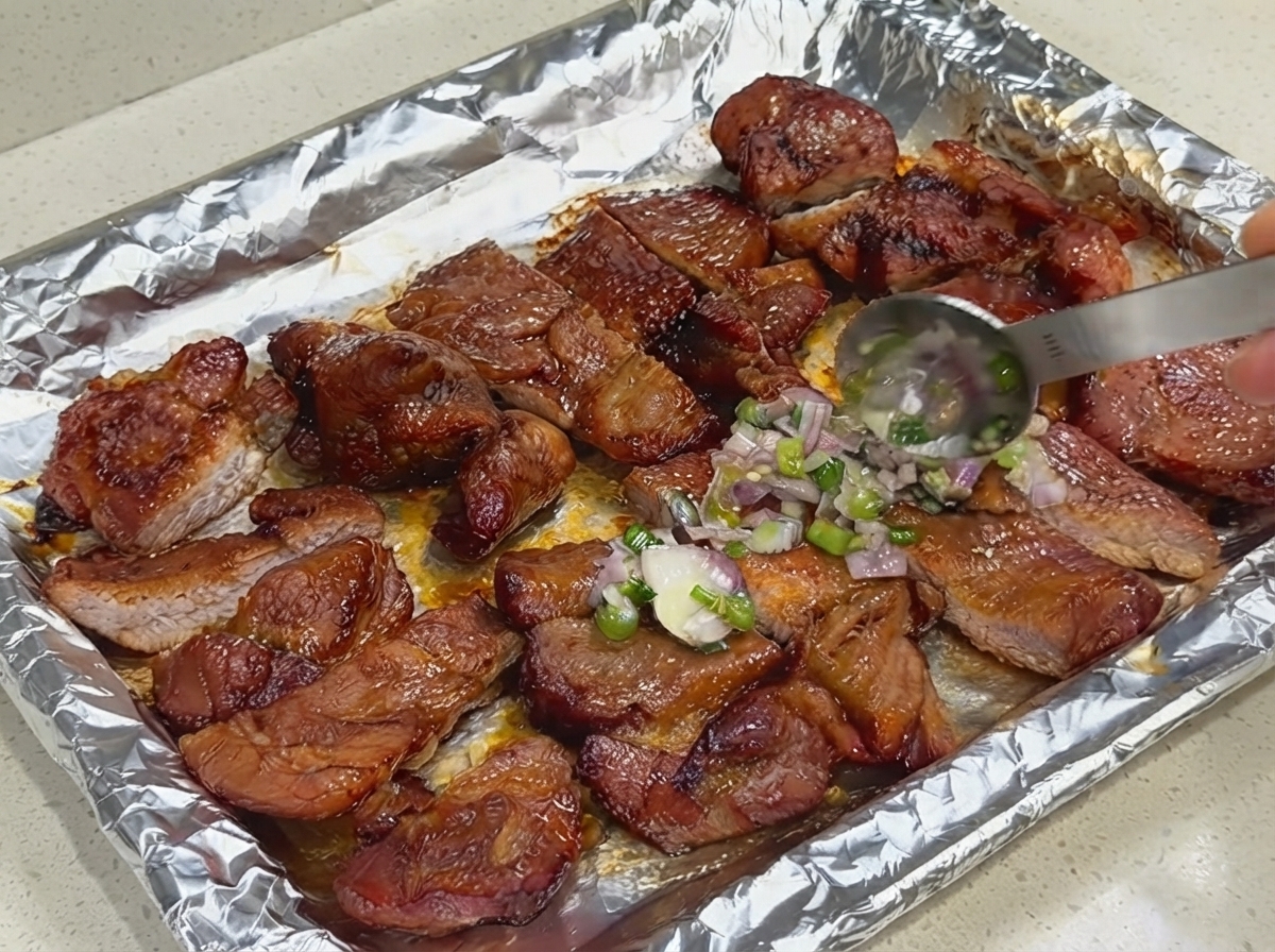 Spoon applying a layer of fresh scallion and shallot sauce onto sliced pork on a foil-lined baking sheet.