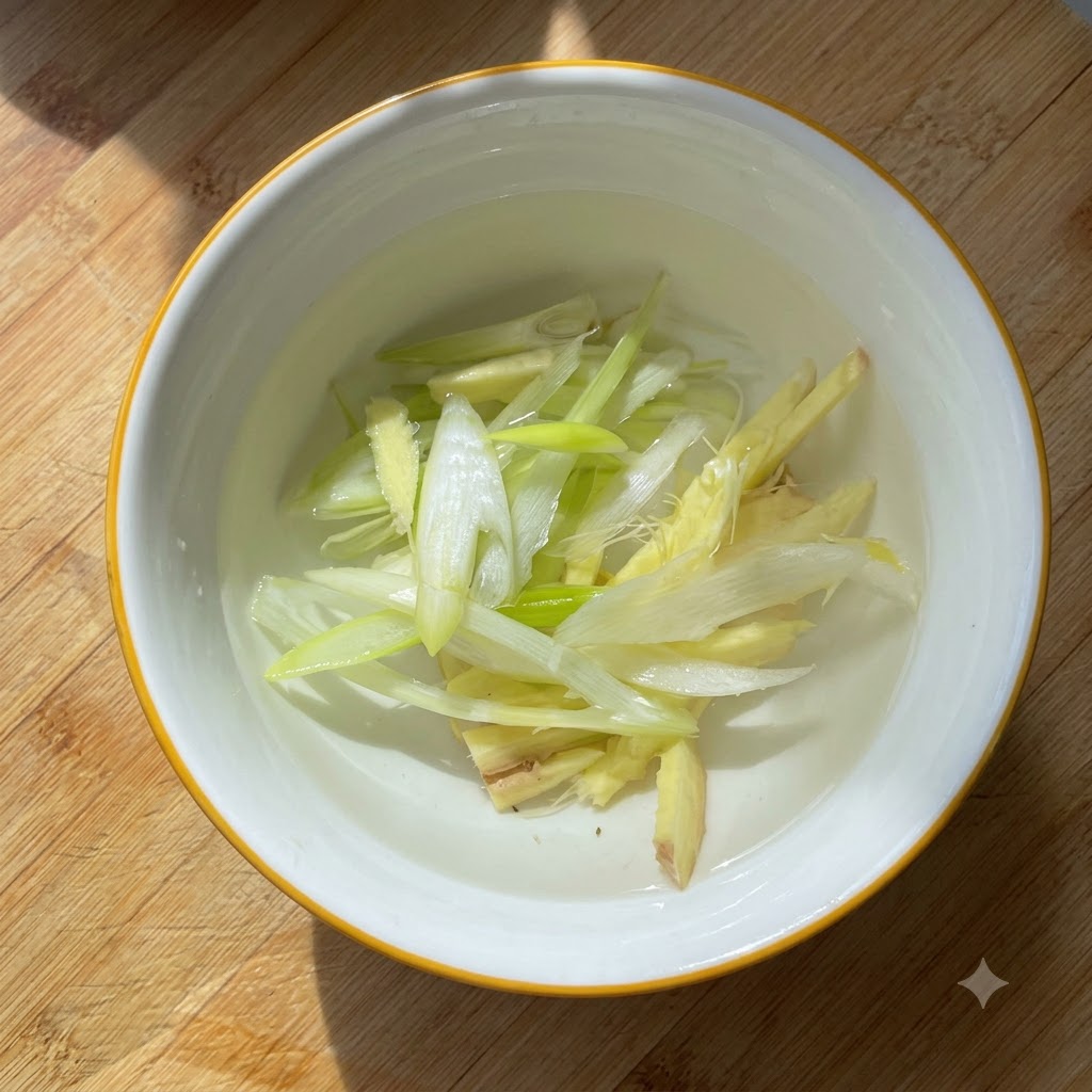 Sliced green onions and ginger pieces steeping in water inside a small white bowl.