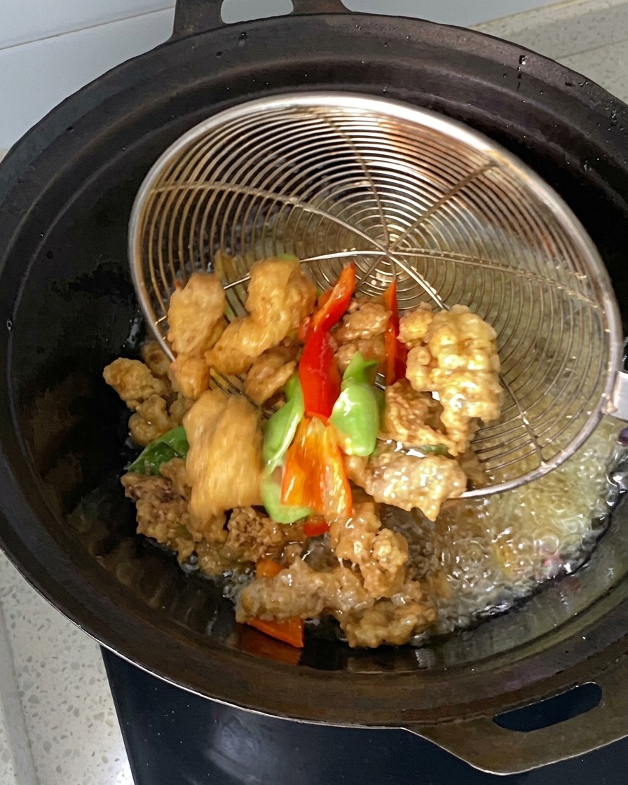 Marinated meat and pepper pieces being lowered and scooped with a wire strainer in a wok of hot oil.