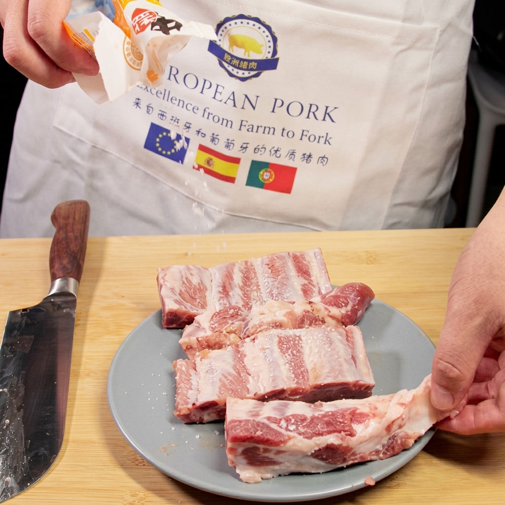 Salt being shaken from a package onto raw pork ribs resting on a plate next to a chefs knife.