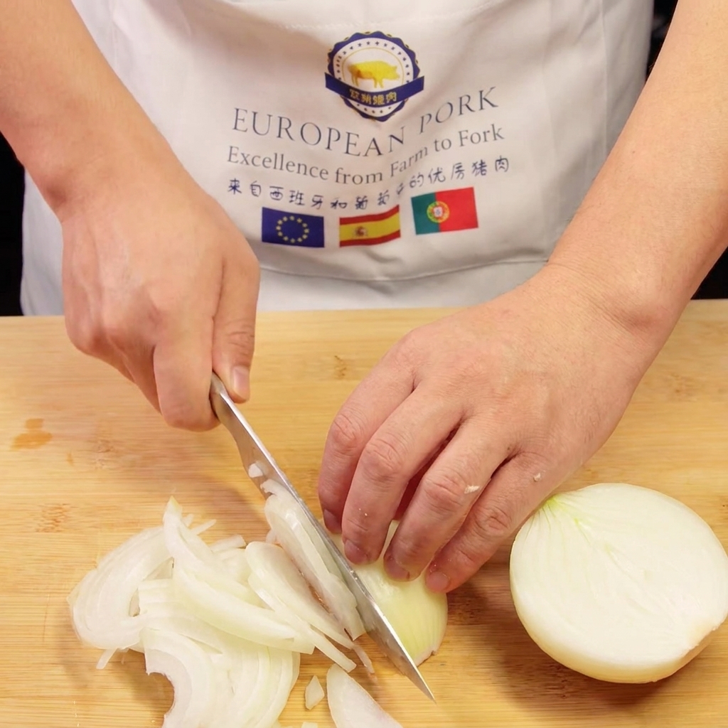 A chefs hands slicing an onion into thin shreds on a wooden cutting board.