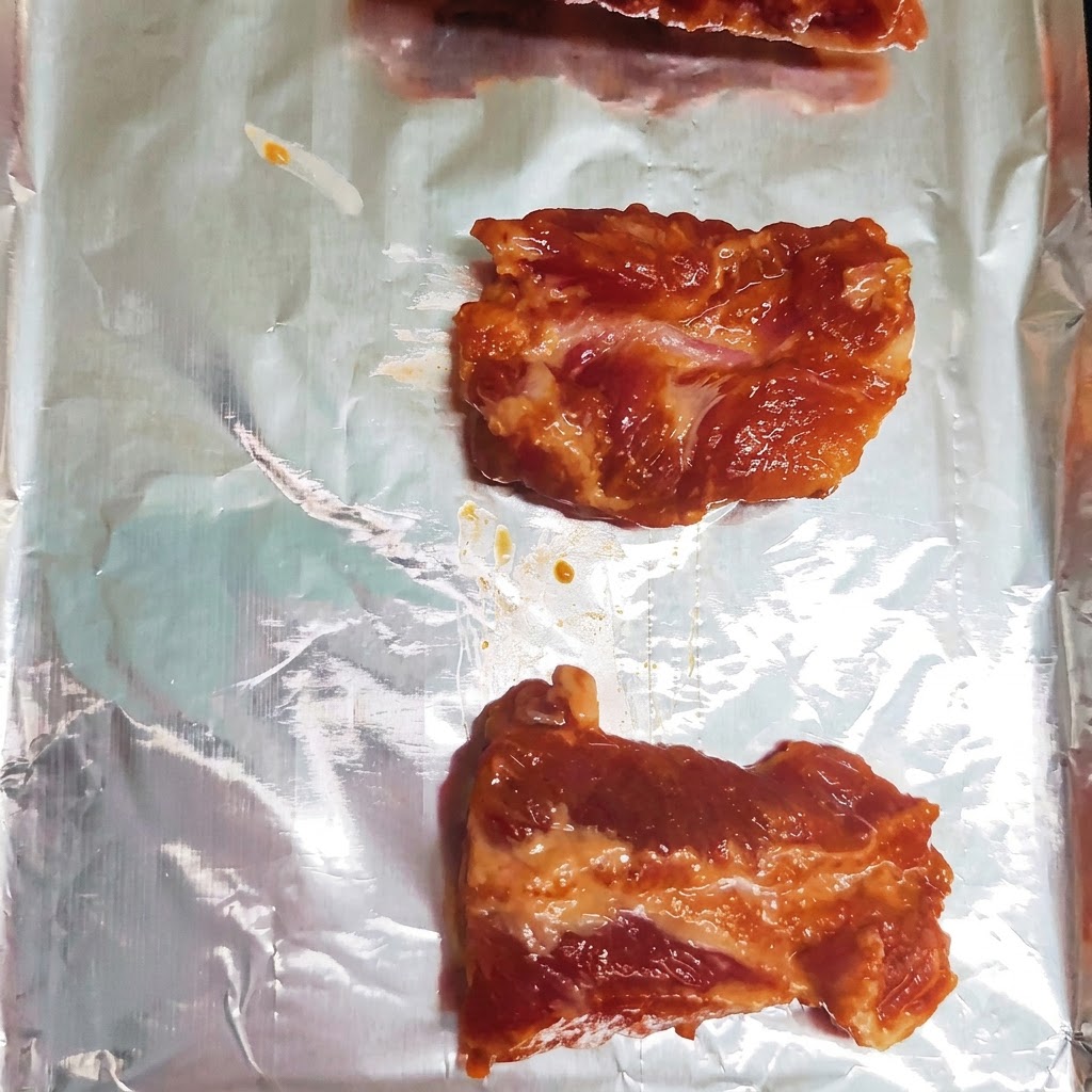Marinated pork rib pieces neatly arranged in a single row on a foil-lined baking sheet.