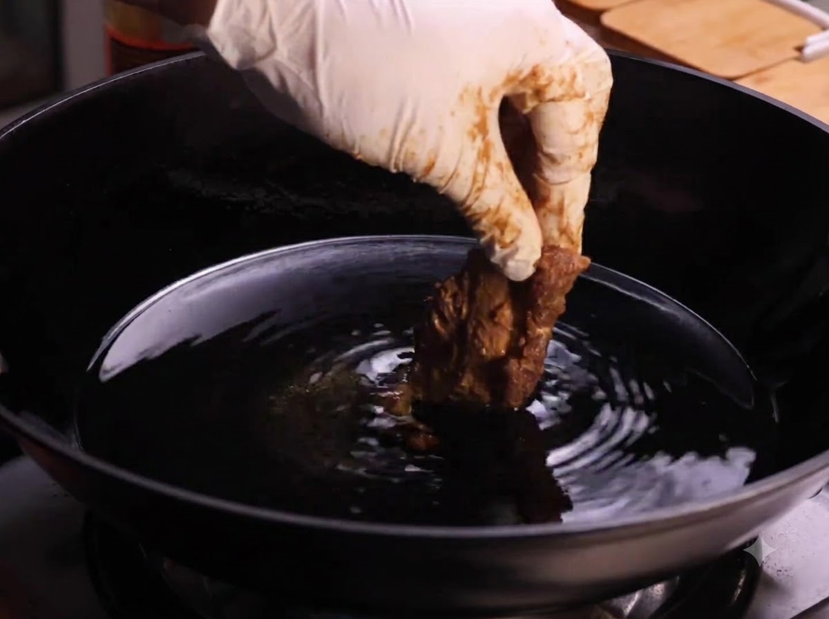A gloved hand lowering a piece of coated pork rib into a wok filled with hot cooking oil.