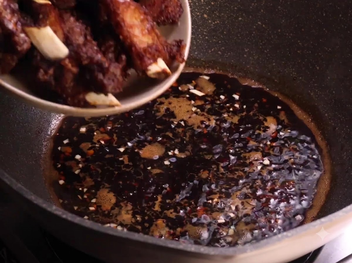 Fried pork ribs being transferred from a plate into the simmering dark sauce in a wok.