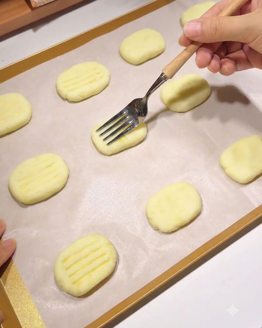 A wooden-handled fork pressing a pattern of parallel lines into a shaped potato pancake on a parchment-lined baking tray.