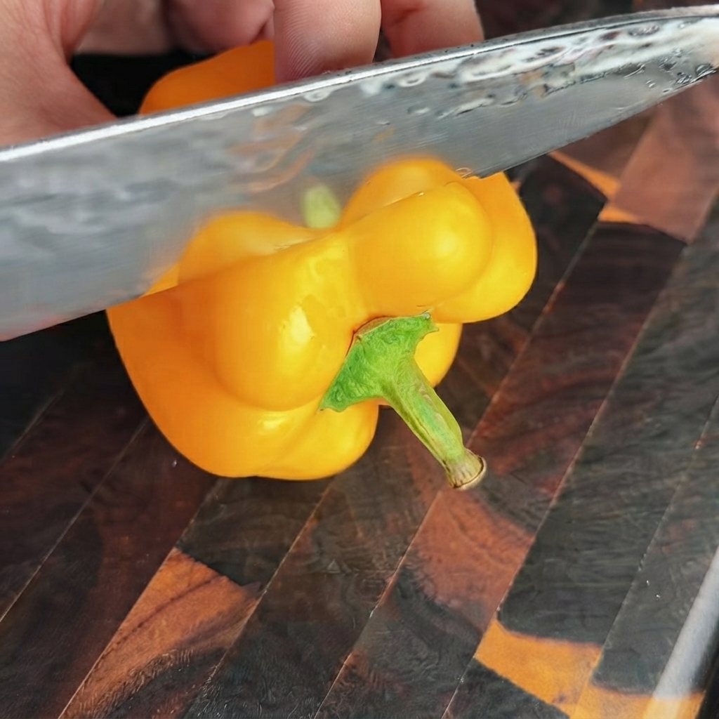A sharp knife slicing off the top section of a bright yellow bell pepper on a wooden cutting board.