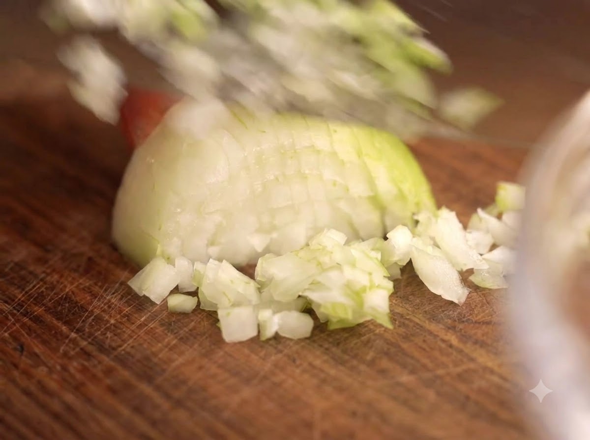 A knife chopping onions and garlic on a wooden board next to a glass bowl of meat.