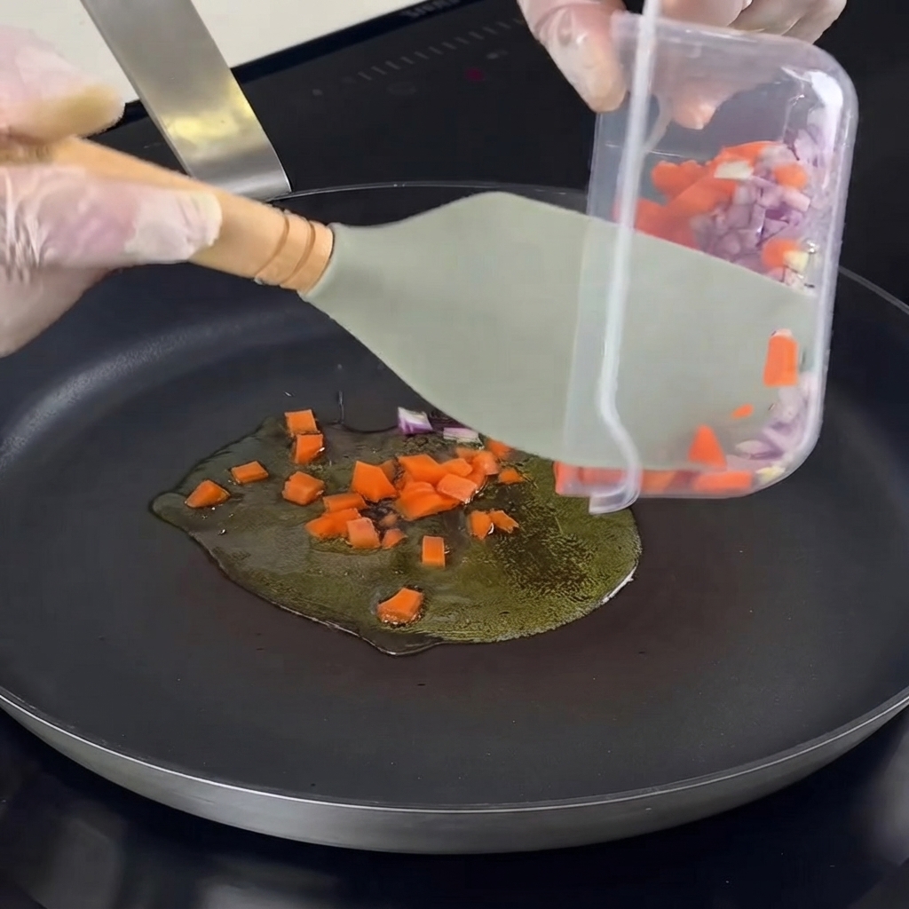 Chopped onions, carrots, garlic, and herbs being transferred into a hot pan with oil.