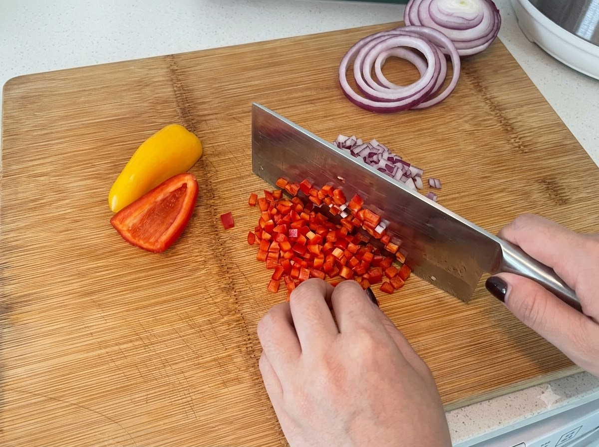Red and yellow bell peppers being diced on a wooden cutting board with a kitchen cleaver.