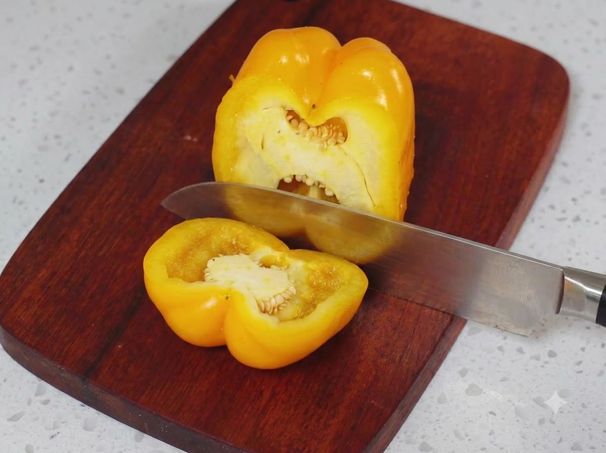 A yellow bell pepper being sliced in half on a wooden cutting board with a kitchen knife.