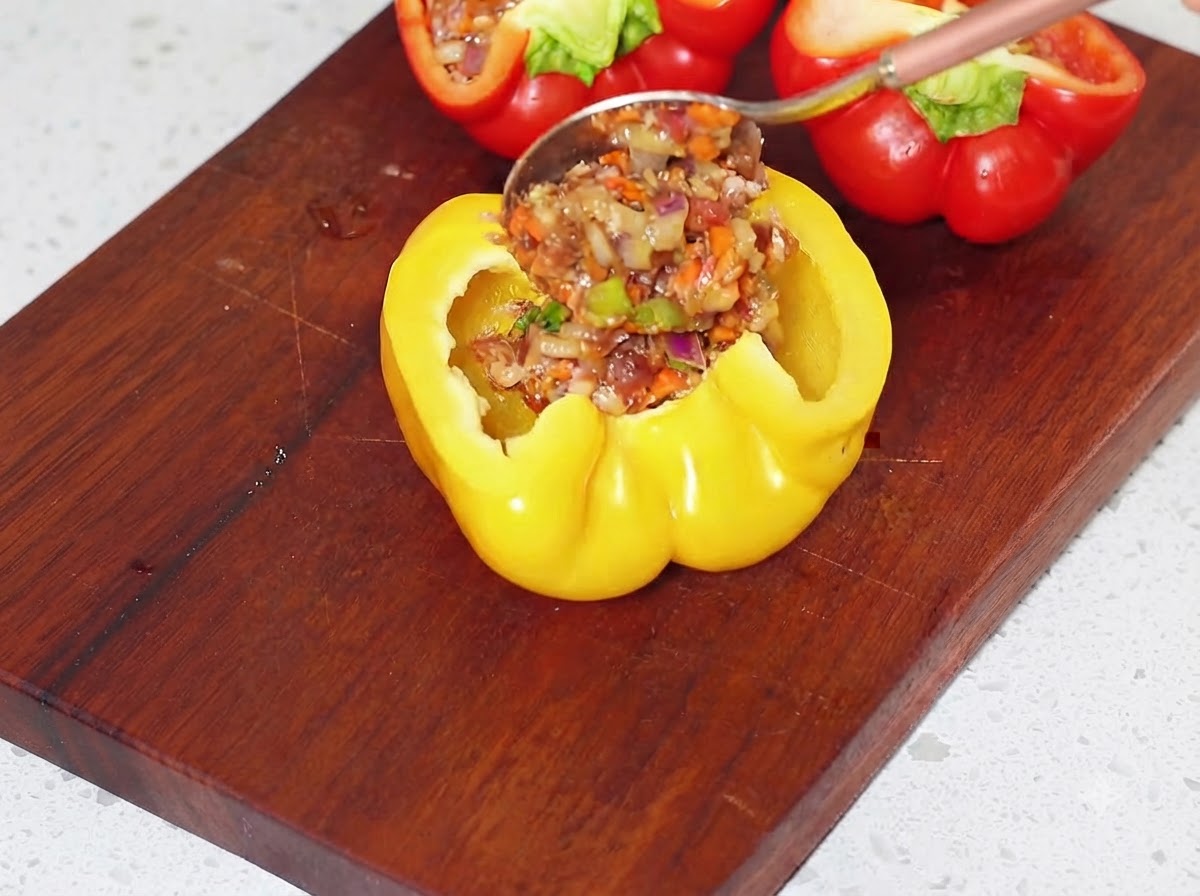 Spoonfully filling the hollowed yellow bell pepper halves with the prepared meat mixture on a cutting board.