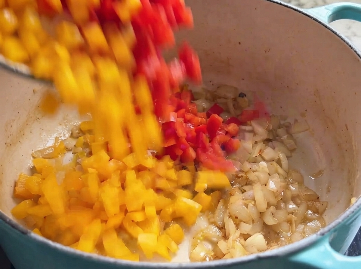 Red and yellow bell pepper dices falling into a cooking pot filled with sauteed onions.