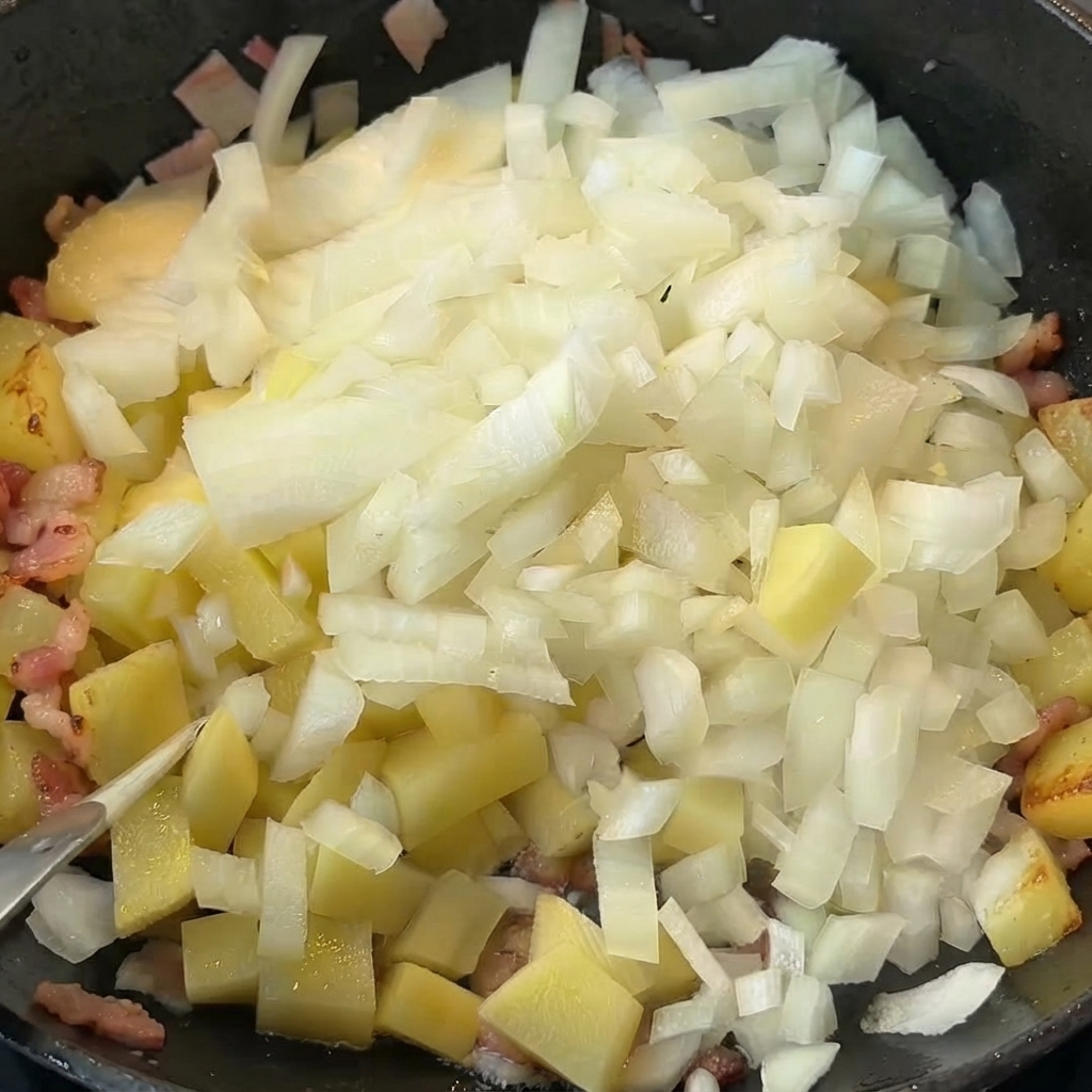 Diced onions being poured on top of cooked potatoes and bacon in a pan.