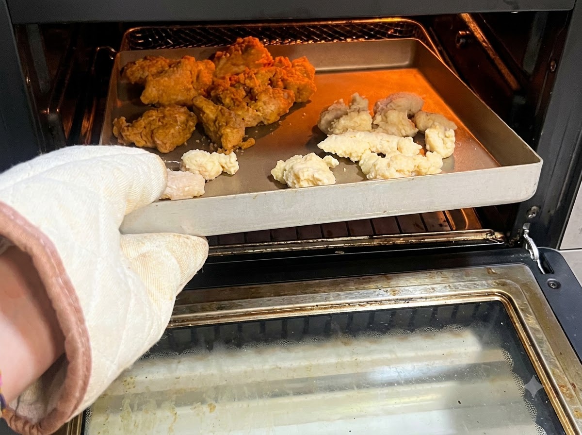 An oven glove holding a tray filled with raw or frozen popcorn chicken and spicy wings inside an air fryer.