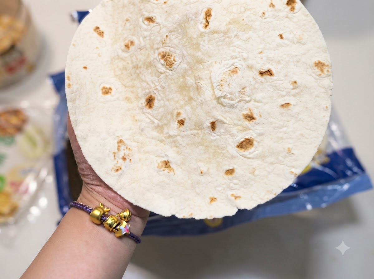 A close-up hand holding a round flour tortilla with characteristic brown spots.