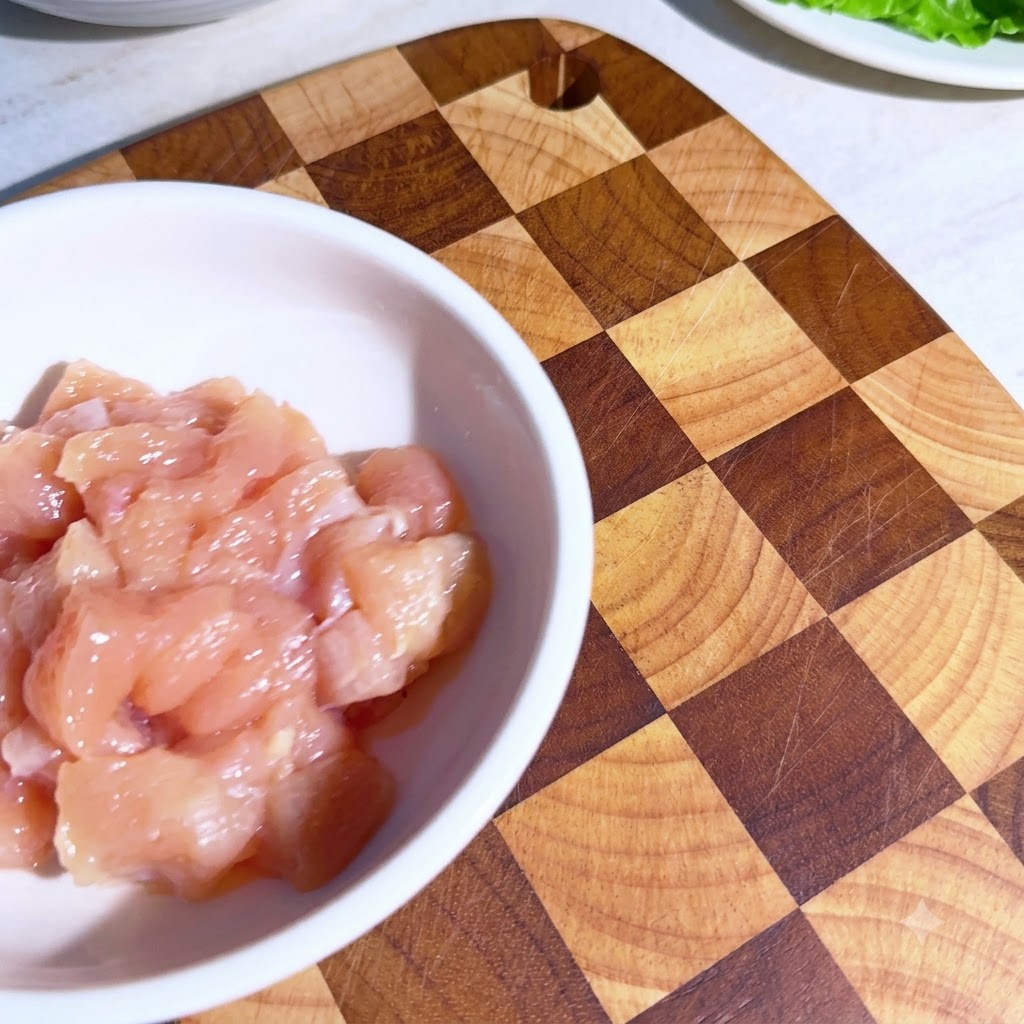 Small chicken pieces in a white bowl with measured spices sprinkled on top before mixing.
