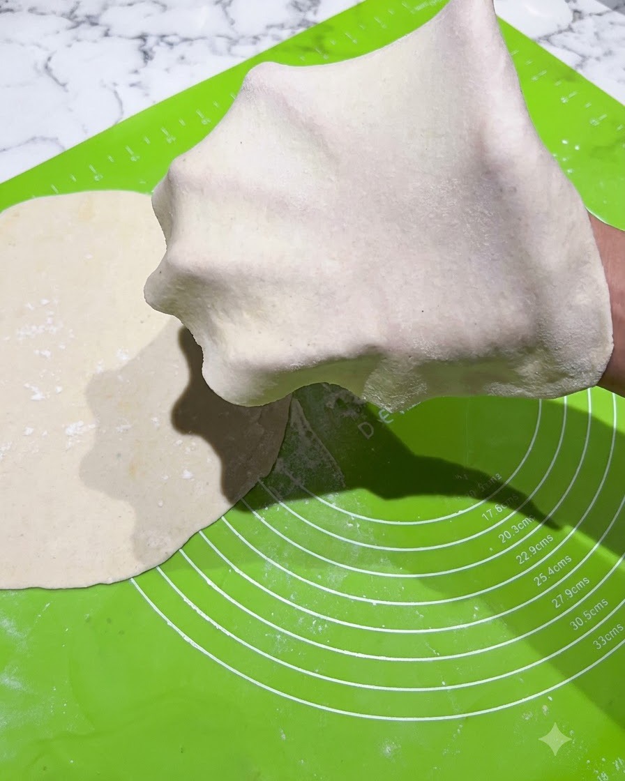 A thin sheet of rolled dough being stretched over a gloved hand on a green mat.