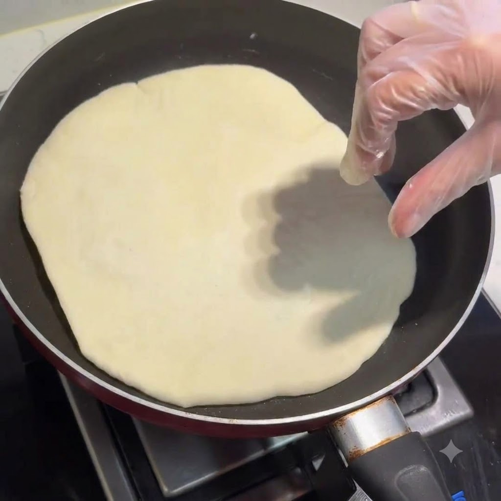 A flatbread cooking in a frying pan with a gloved hand adjusting it.