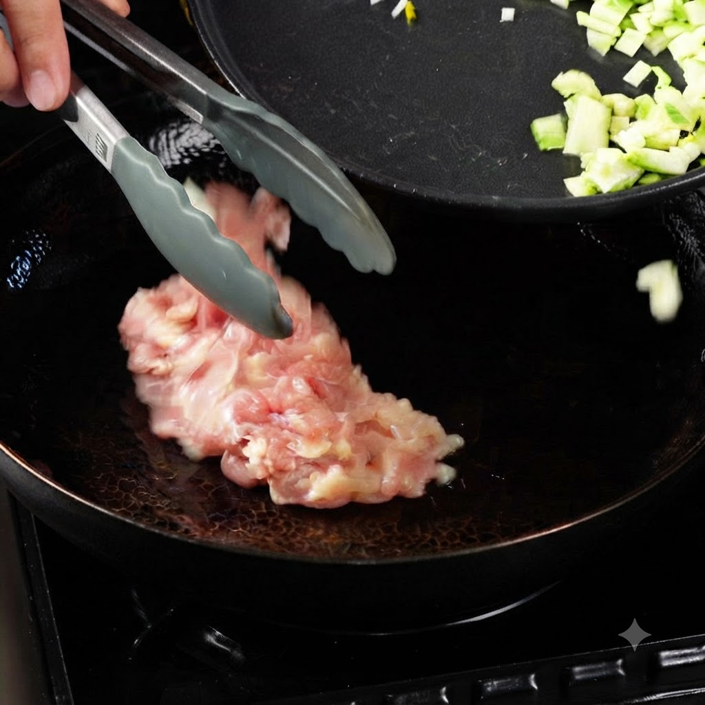 Tongs placing raw chicken pieces into a heated wok with oil, next to a pan containing diced vegetables.