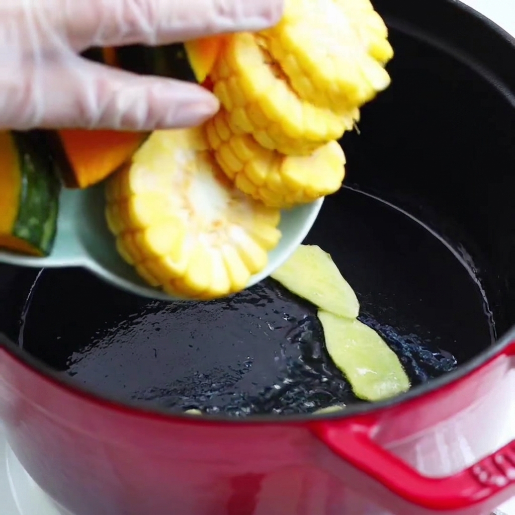 Pumpkin chunks and corn cobs being poured from a dish into the red pot.