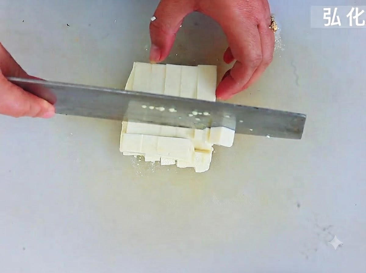 Soft tofu being sliced into uniform square cubes with a large cleaver on a cutting board.